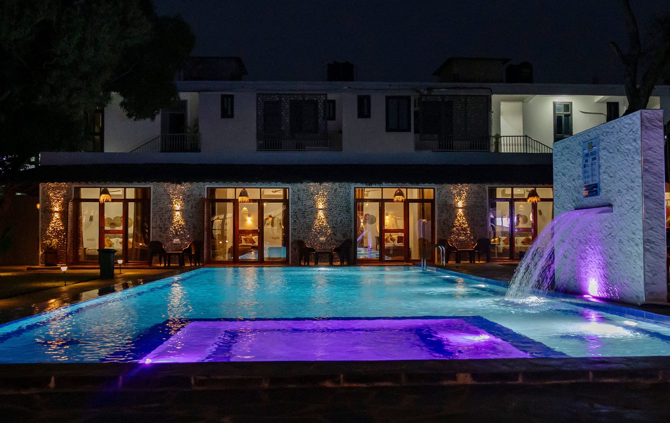 The pool lit turquoise and violet after dark, warm light in the room doorways