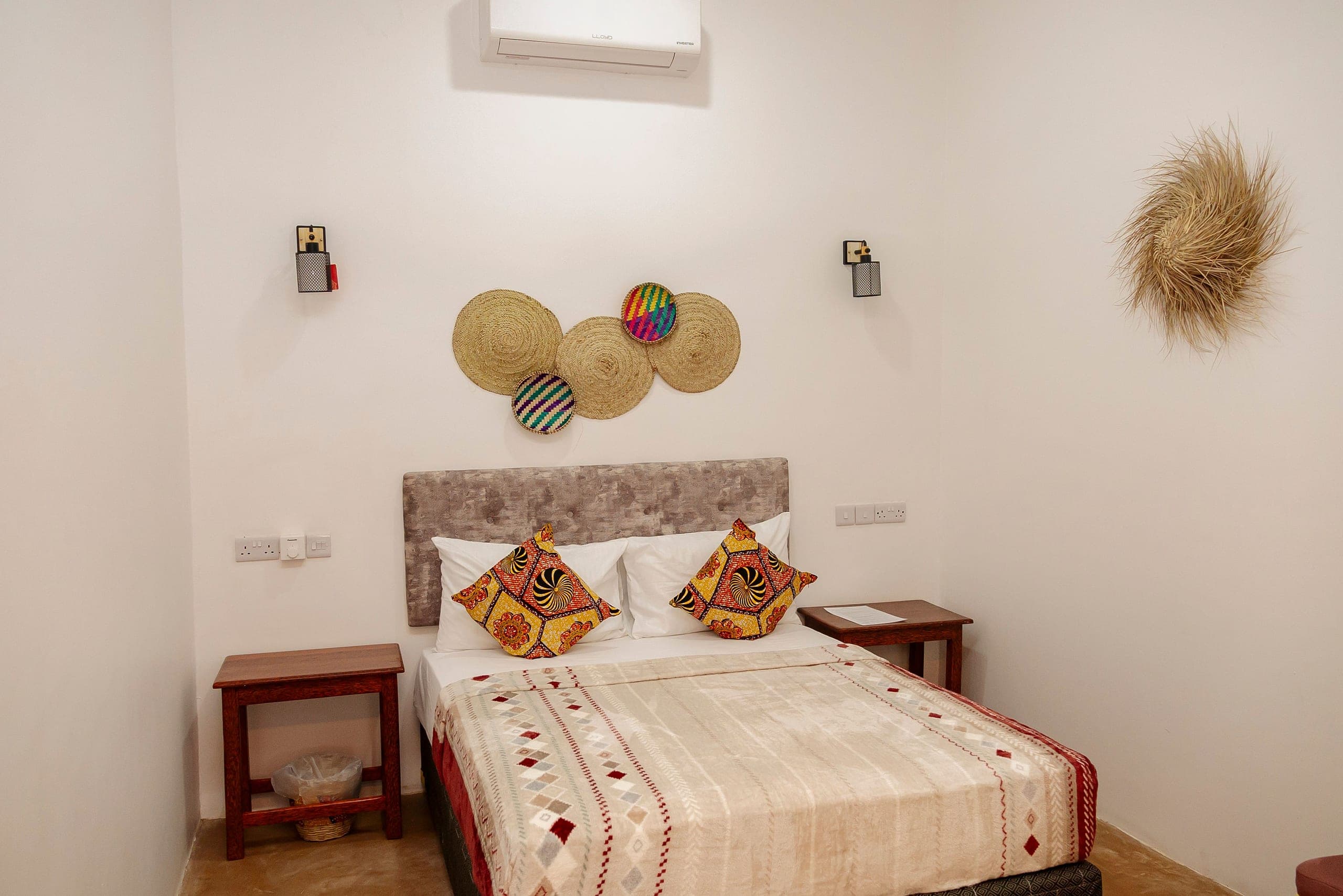Double room with a soft grey headboard, kanga cushions and woven wall discs